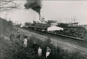 Japanese men and women in dresses and suits stand on a hill. Railroad tracks tall ships and steam from a factory building in the background